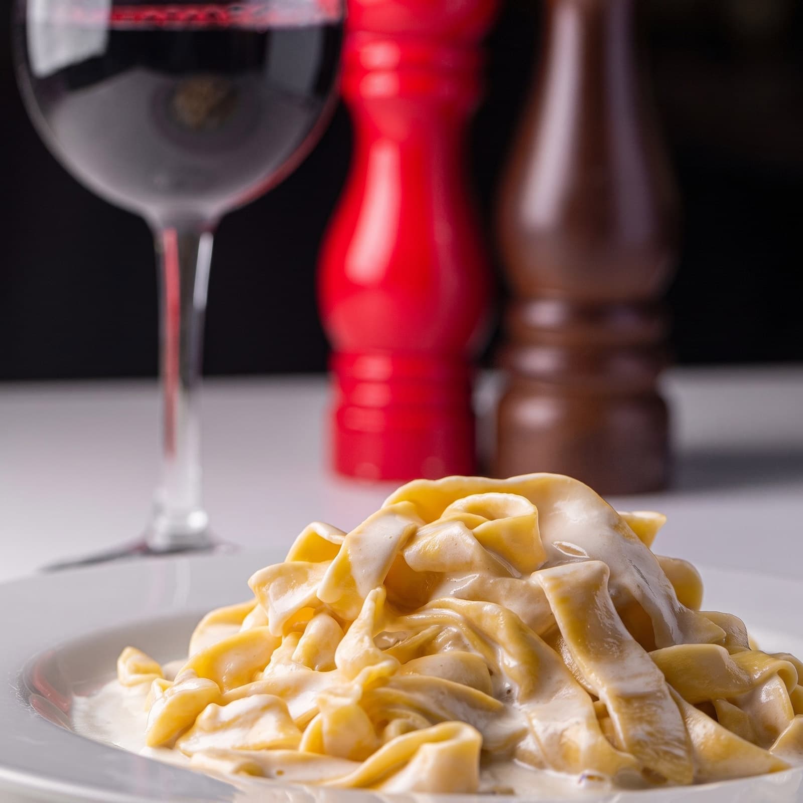 A creamy pasta dish plated on a white restaurant table, with a glass of red wine and table condiments in the background.